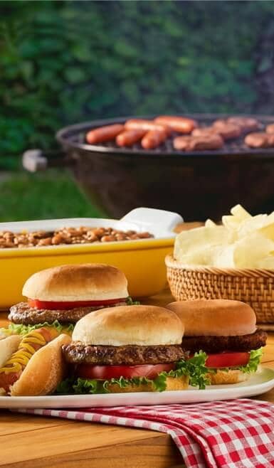A picnic table with hamburgers, hot dogs, potato chips, and baked beans. A grill with hot dogs and hamburgers is in the background.